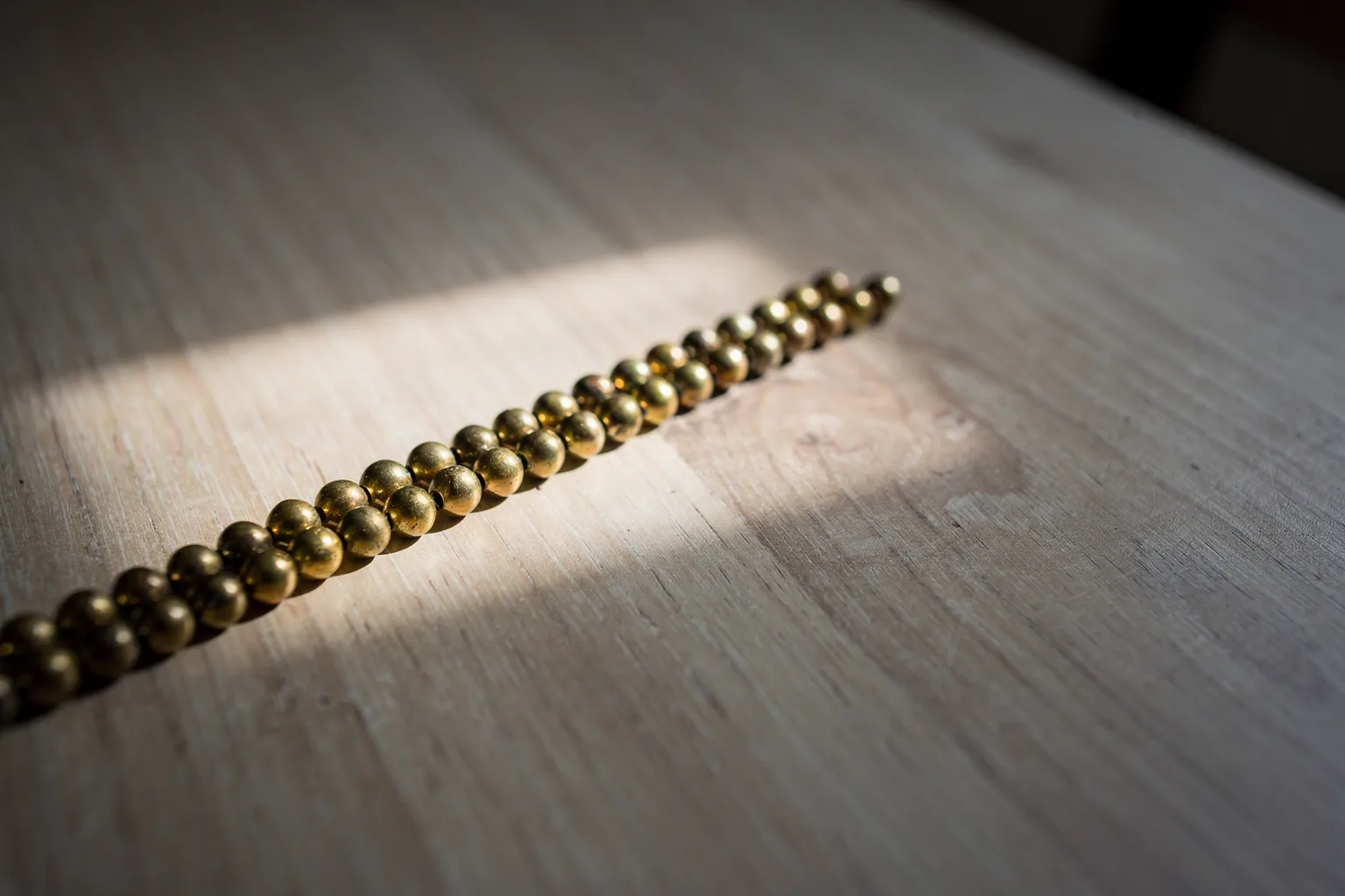 A row of brass beads laid diagonally across a light wooden table, crossed by a band of window light: a few beads are lit, the rest stay in shadow.