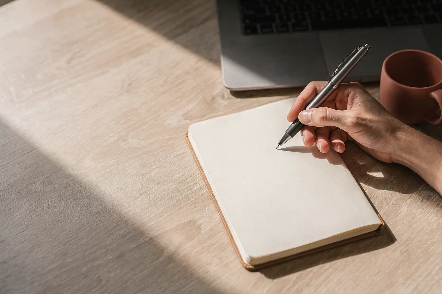 A hand holds a pen above the blank page of an open notebook on a light wood desk, next to a closed laptop and a terracotta mug, in low morning light.