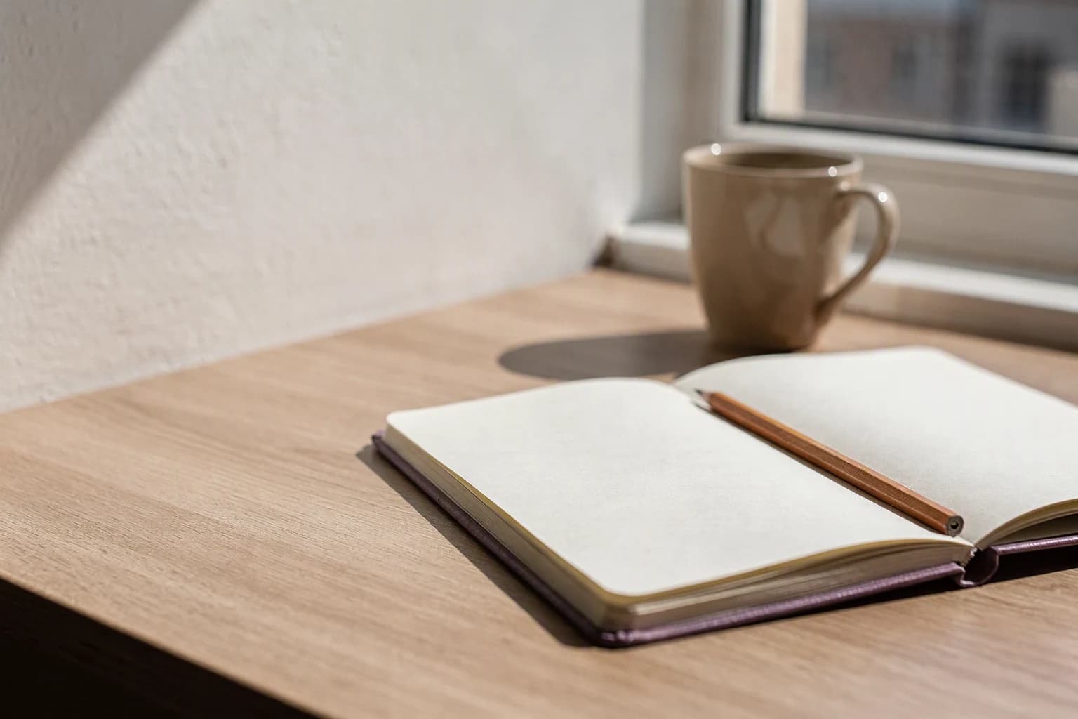 An open notebook on a blank page, a wooden pencil resting across it, on a light wood desk by a window, with a beige ceramic mug in the background.